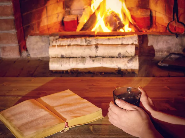 Les mains des femmes se réchauffent sur une tasse de thé chaude près d'une cheminée en feu, un livre ouvert est sur la table — Photo