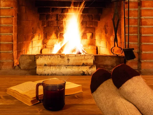 Tee, Buch, Frauenfüße in warmen Socken auf einem Holztisch vor einem brennenden Kamin — Stockfoto