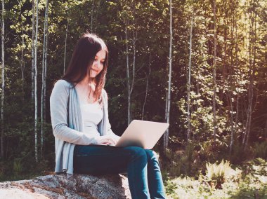 Genç ve güzel bir kız ormanda büyük bir kayanın üzerinde otururken dizüstü bilgisayarla çalışıyor. Serbest konsept