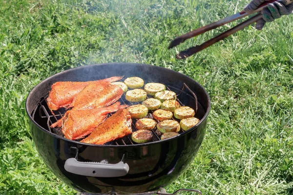 Piknik na świeżym powietrzu w lecie. Gotowanie na okrągłym grillu ryb morskich bas i cukinia plastry — Zdjęcie stockowe