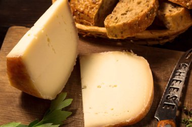 Bir dilim sert peynir, maydanoz, kekik, peynir bıçağı, ahşap masada tahıl ekmeği kesme