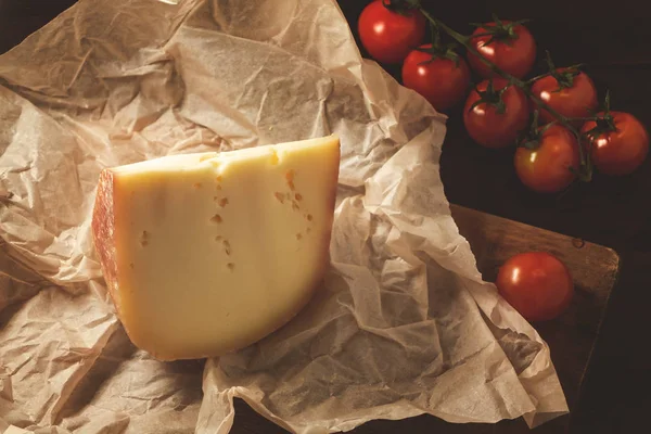 Stuk harde kaas op ambachtelijk papier en cherry tomaten op een houten tafel — Stockfoto