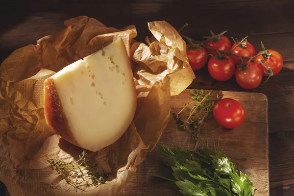 Stuk harde kaas op kraftpapier, peterselie, tijm, en cherry tomaten op een houten tafel — Stockfoto