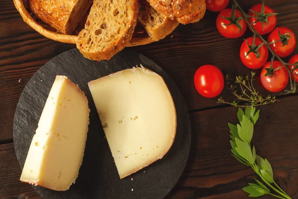 Trozos de queso duro en plato de pizarra, perejil, tomillo, pan de grano y tomates cherry en una mesa de madera, vista superior, puesta plana — Foto de Stock