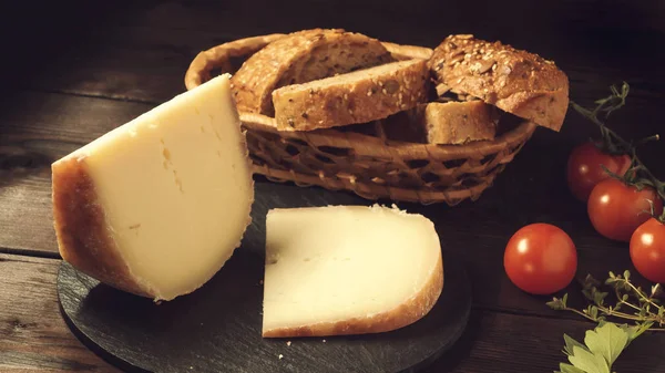 Trozos de queso duro en plato de pizarra, perejil, tomillo, pan de grano y tomates cherry en una mesa de madera — Foto de Stock