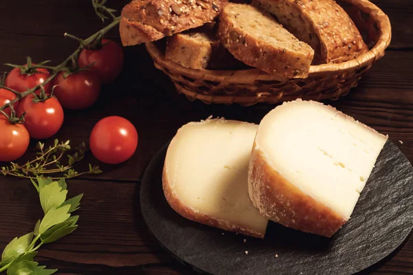 Trozos de queso duro en plato de pizarra, perejil, tomillo, pan de grano y tomates cherry en una mesa de madera — Foto de Stock