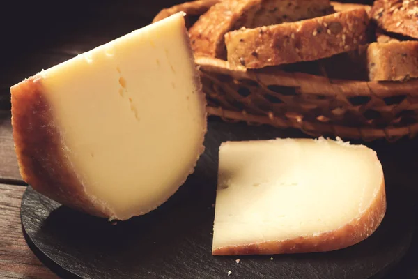 Trozos de queso duro en plato de pizarra, pan de grano en una mesa de madera — Foto de Stock