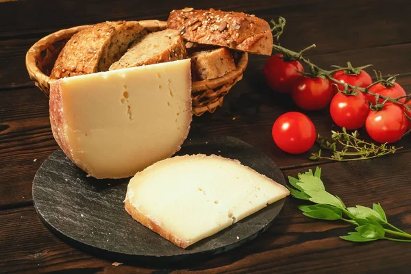 Kousky tvrdého sýra na břidlicové misce, petržel, Thyme, obilný chléb a třešňová rajčata na dřevěném stole — Stock fotografie