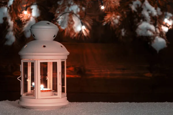 Lantaarn met een brandende kaars op de sneeuw tegen de achtergrond van een oude houten muur versierd met kerstboom takken en kerstverlichting. Ruimte kopiëren voor tekst — Stockfoto