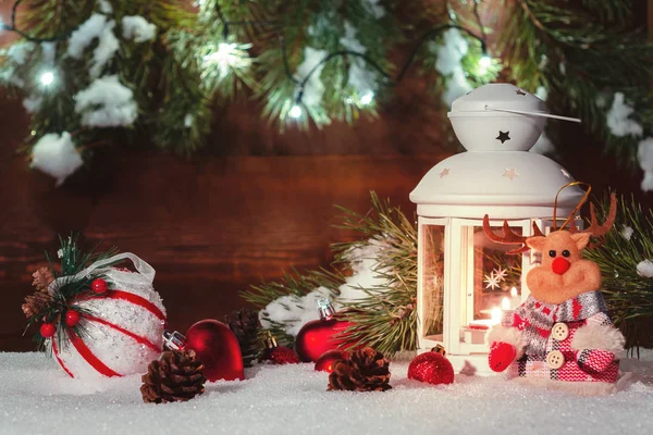 Lanterna branca com uma vela acesa fica na neve cercada por decorações de Natal no fundo de uma parede de madeira, galhos de árvore de Natal e luzes — Fotografia de Stock