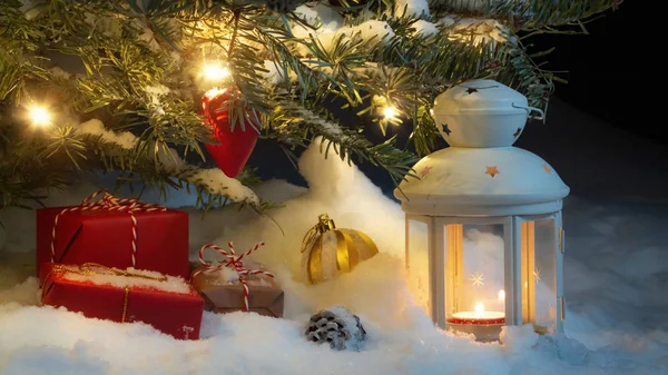 Tarjeta de Navidad - regalos y una linterna en la nieve bajo un árbol de Navidad decorado con luces y decoraciones de Navidad — Foto de Stock