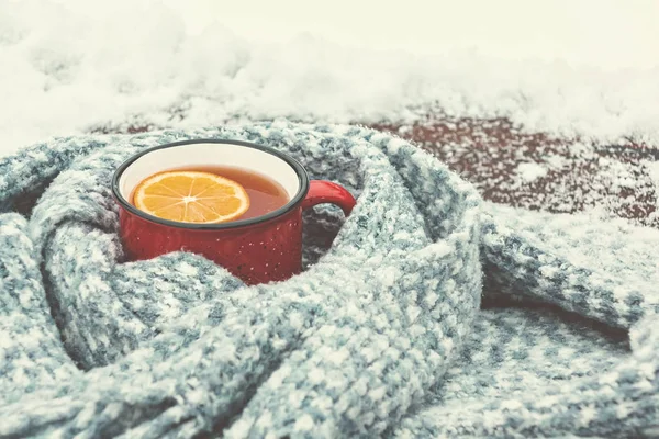 Red enameled cup of hot tea with lemon wrapped in a knitted scarf on a snowy wood table — Stok Foto