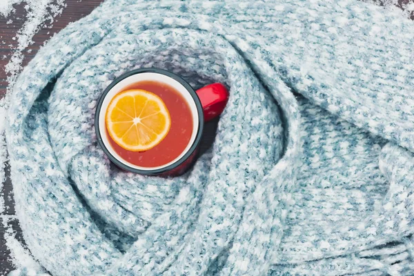 Red enameled cup of hot tea with lemon wrapped in a knitted scarf on a snowy wooden table, top veiw, flat lay