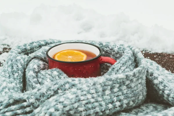 Red enameled cup of hot tea with lemon wrapped in a knitted scarf on a snowy wooden table