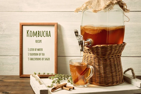 Fresh homemade kombucha fermented tea drink in a jar with faucet and in a cup on a white tray, the recipe for cooking in the frame, wooden background