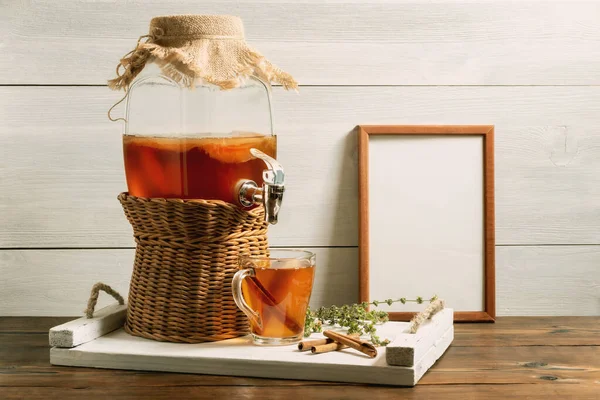 Fresh homemade kombucha fermented tea drink in a jar with faucet and in a cup on a white tray, frame with copyspace, wooden background