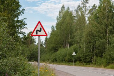 Kırmızı çerçeveli üçgen beyaz yol işareti. Asfalt bir yolun kenarında DANGEROUS DURN