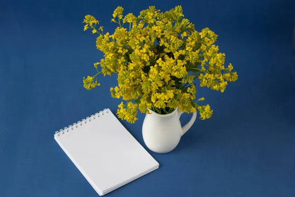 Bouquet of wild flowers in a vase on a blue background, top view . Fresh spring flowers. Blank notebook with with space for text.