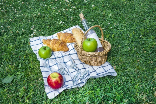 Romantic summer picnic lunch outdoors. Wicker basket with croissants, apples and bottle of rose wine  on checkered cloth on grass in park. opy space for text