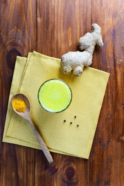 Golden milk from turmeric, vegetable milk, pepper, ginger, coconut oil, maple syrup in a glass on a wooden table. Traditional indian drink, top view.