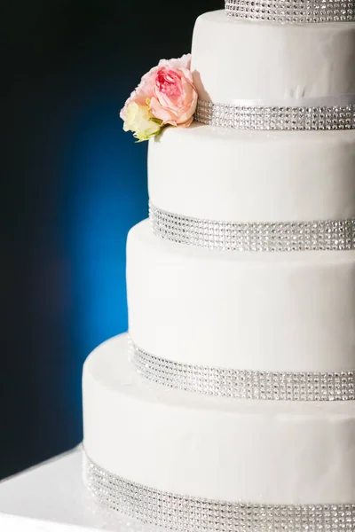 A four-tiered cake with white cream silver rhinestones and live roses stands on a blue background