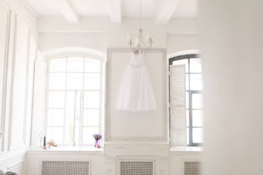 Çerçeve içinde iki pencere arasında duvarda beyaz bir odada muhteşem bir gelinlik asılı. Pencere nin üzerinde bir buket çiçek var.