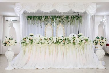 Gelin ve damat için şık düğün prezidonu. Beyaz bir masa örtüsü, büyük gül buketleri ile masa. Kenarları boyunca çiçekler ve at kafaları duvara asılı zemin vazolar vardır. Altın oyma sırtlı sandalyeler.