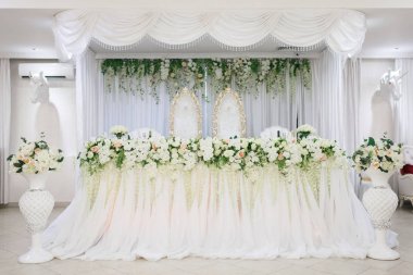 Chic wedding presidium for the bride and groom. Table with a white tablecloth, large bouquets of roses. Along the edges there are floor vases with flowers and horses' heads hang on the wall. Chairs with gold carved backs. clipart