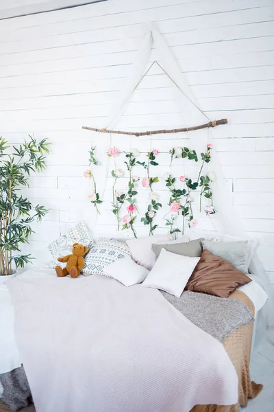 In a bright room there is a bed with a lot of multi-colored pillows on it and a teddy bear. At the headboard hangs a panel of pink and white roses. Nearby is a houseplant.