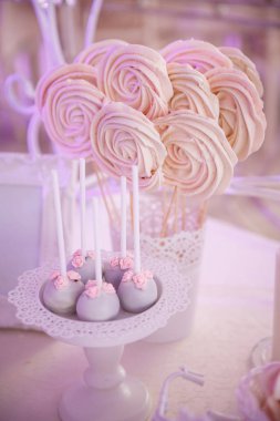 Candi bar. Ahşap şişler üzerinde el yapımı pembe marshmallow dantel kenarları ile beyaz bir cam duruyor. Yakınlarda bir vazoda küçük çiçekler ve çubuklar içeren kek topları bulunmaktadır. Bulanık arka plan.