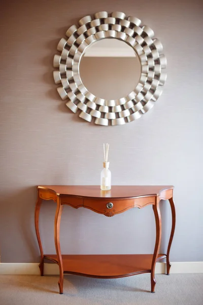 On the gray wall hangs a round mirror in a carved frame. Raise is a wooden table with curved legs. On the tabletop bottle with incense sticks.