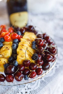 Masada beyaz dantel masa örtüsü, bordo ve kırmızı kiraz meyveleri, böğürtlen, yaban mersini ve sarı sulu ananas ile bir cam tabak.