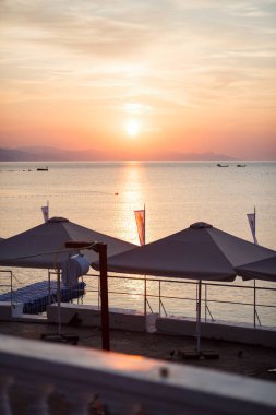 Siyah deniz üzerinde pembe gündoğumu. Suyun üzerinde güzel bir ışık yolu. Beach. Ön planda güneşten gelen tenteler. Mesafe tekneler.