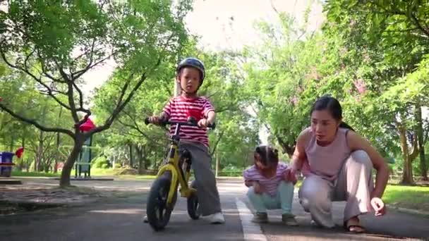 Die asiatische Familienmutter und ihre Kinder spielen Wettkampflauf und Fahrradfahren auf der asphaltierten Straße im Park. Gemeinsam Spaß draußen haben — Stockvideo