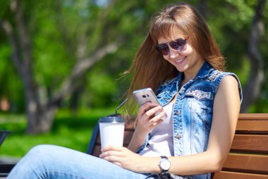 Güneşli bir dayteenager üzerinde bir bankta bir parkta otururken, on-line Alışveriş kavramı bir akıllı telefon ile güzel neşeli kız.