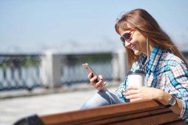 Genç mutlu kadın bir akıllı telefon kullanıyor. O bir kağıt bardak kahve tutuyor
