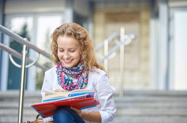 Üniversite ya da üniversitenin merdivenlerinde kitap yazıp okuyan gülümseyen genç bayan sınavlara hazırlanıyor. Eğitim kavramı, kopyalama alanı