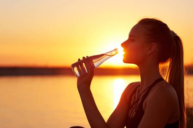 Siluet kadın koşudan sonra şişeden su içiyor ya da sahilde yoga yapıyor. Gün batımında spor ve rahatlama konsepti.