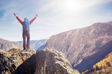 Sırt çantalı bir yürüyüşçü dağın tepesinde duruyor, elleri havada ve güneşin doğuşunun keyfini çıkarıyor.