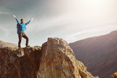 Sırt çantalı bir yürüyüşçü dağın tepesinde duruyor, elleri havada ve güneşin doğuşunun keyfini çıkarıyor.