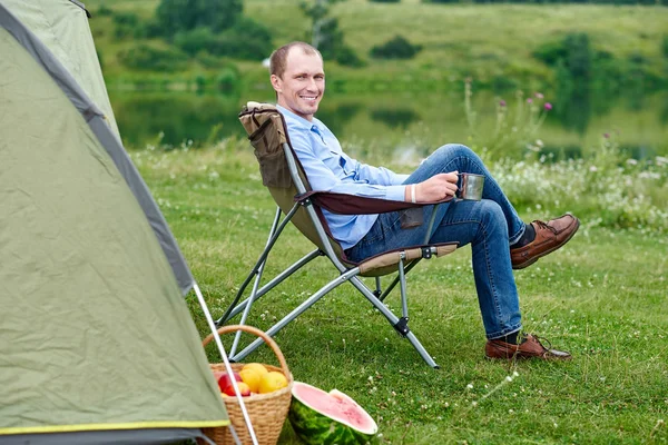 Giovane freelance seduto sulla sedia e rilassante davanti alla tenda in campeggio nella foresta o prato. Attività all'aperto in estate. Avventura in viaggio nel parco nazionale. tempo libero, vacanza, relax — Foto Stock