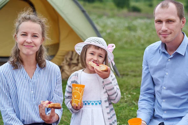 Famiglia felice al picnic in campeggio. Madre, padre e figlia mangiano vicino a una tenda nel prato o nel parco — Foto Stock