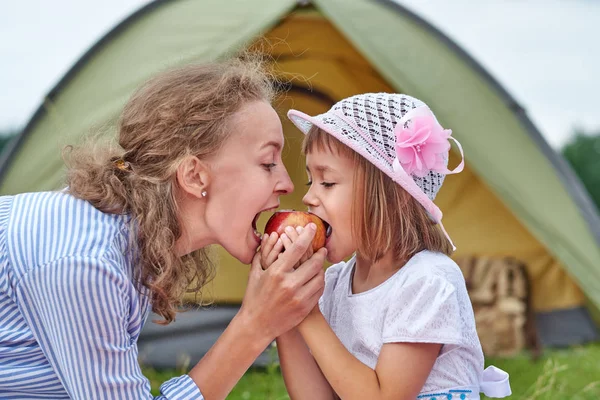 Anne ve kızı çayırda ya da parkta bir çadırın yanında elma yiyorlar. Mutlu aile kampta piknik yapıyor. — Stok fotoğraf