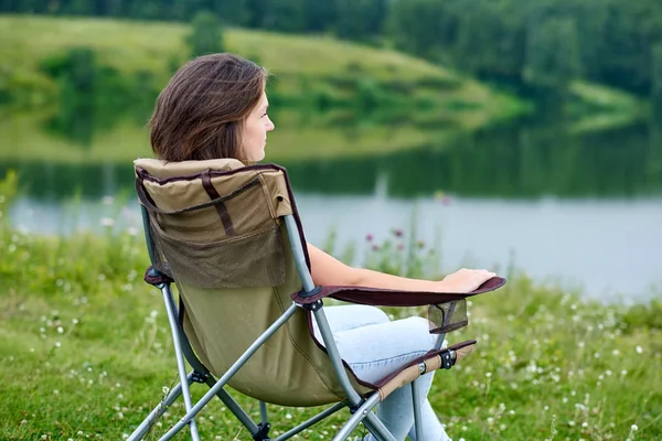 Sandalyede oturan ve gölün yanındaki doğada dinlenen genç bir kadın. Yazın açık hava aktivitesi. Ulusal parkta maceraya atılmak. Boş zaman, tatil, rahatlama. — Stok fotoğraf