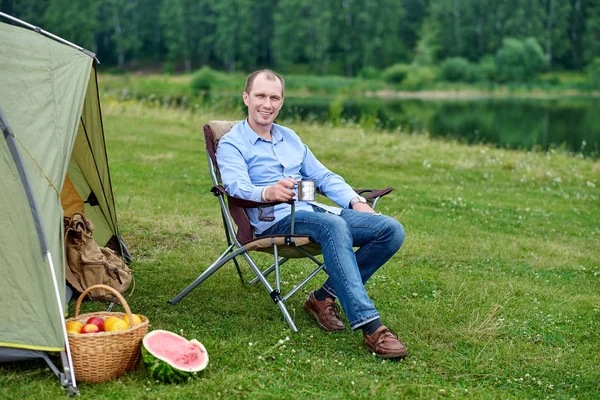 Joven freelancer sentado en la silla y relajarse frente a la tienda de campaña en el camping en el bosque o prado. Actividad al aire libre en verano. Aventura viajando en el parque nacional. ocio, vacaciones, relajación — Foto de Stock
