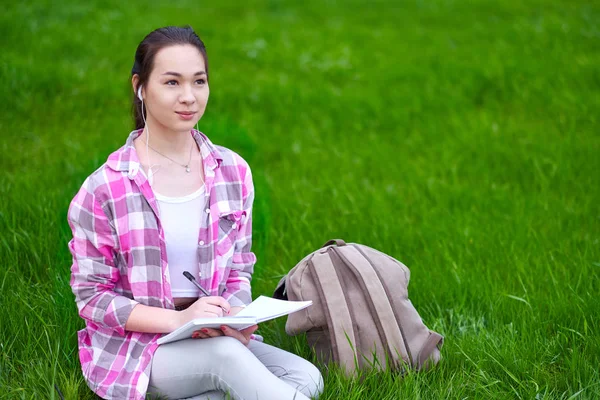 Jeune femme asiatique écrit dans un cahier assis sur la prairie d'herbe — Photo
