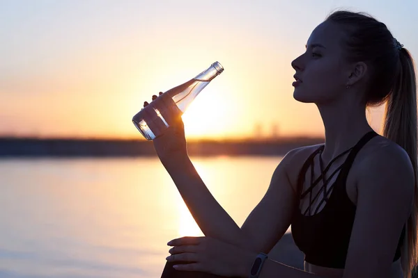 Mujer silueta bebiendo agua de botella después de correr o yoga en la playa. Perfil femenino de fitness al atardecer, concepto de deporte y relajación —  Fotos de Stock