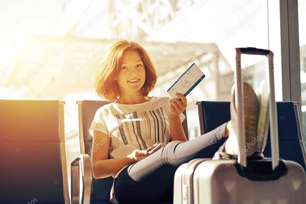 Smiling woman with ticket and baggage in airport. Air travel concept with young casual girl sitting with hand luggage suitcase at gate waiting in terminal.