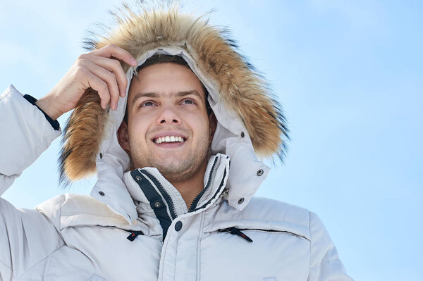 Fashion young beautiful man in a white jacket with fur hood in winter