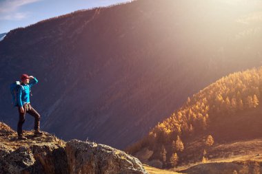 Sırt çantalı bir yürüyüşçü dağın tepesinde duruyor, elleri havada ve güneşin doğuşunun keyfini çıkarıyor.
